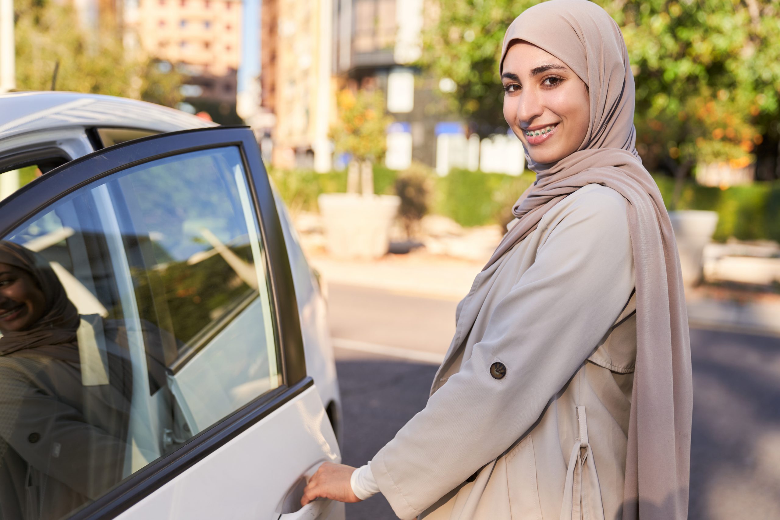 A woman in a tan coat and a scarf is standing next to a car door. She is smiling and she is happy